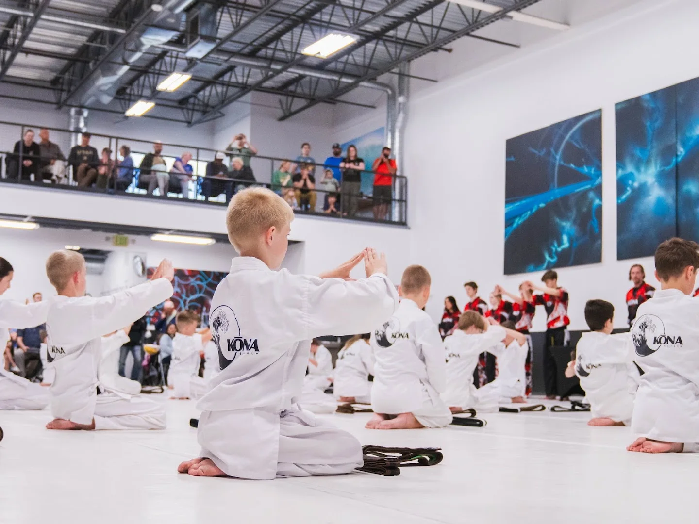 Students kneeling in rows during a promotion event, with families watching from the balcony at Kona Karate